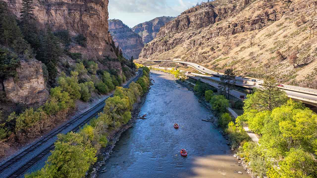 Two orange whitewater rafts float down the Colorado River in the Glenwood Canyon. I-70 is visible on the right and a railroad is left of the river.