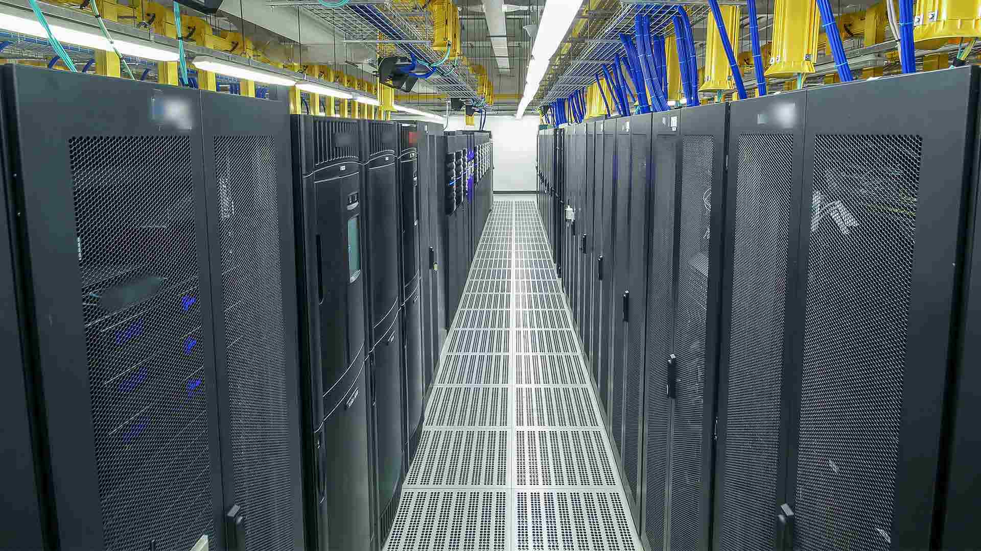 Two rows of tall computer servers a with yellow and blue wires emerging from the top of them routed to conduit in the ceiling.