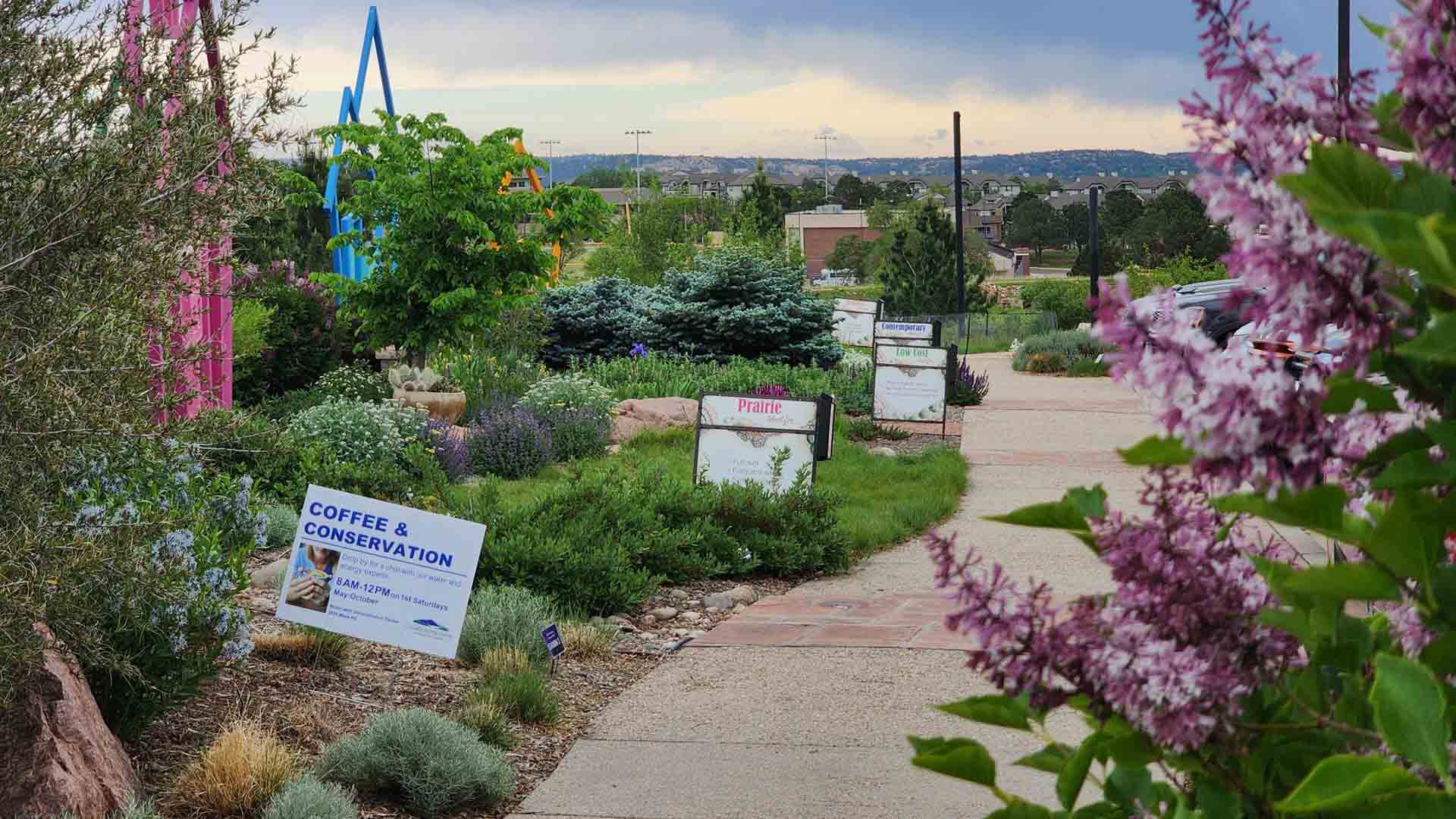 Green varied landscapes next to a concrete sidewalk. There are signs labeling the different demonstration landscapes and their layout. Another sign has visible text that reads 