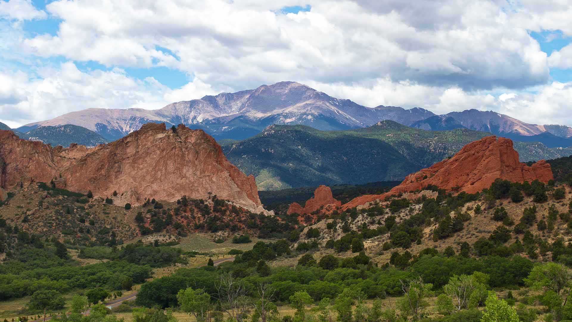 Pikes Peak towers behind the red rock formations of Garden of the Gods on a partly cloudy day.