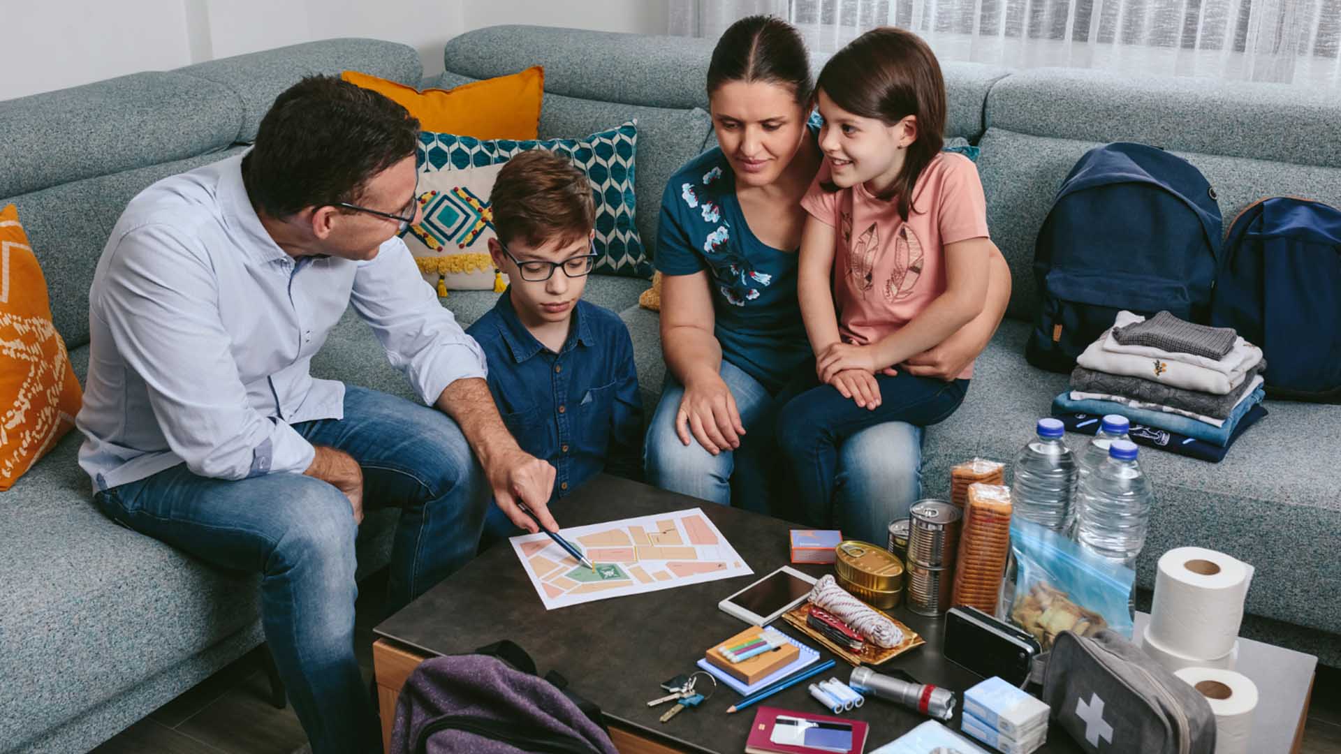 A family of four prepares for an emergency in their living room. The father points to a map on the coffee table, surrounded by emergency supplies like canned food, water, a first-aid kit, and backpacks. The mother embraces their daughter, while their son watches intently.