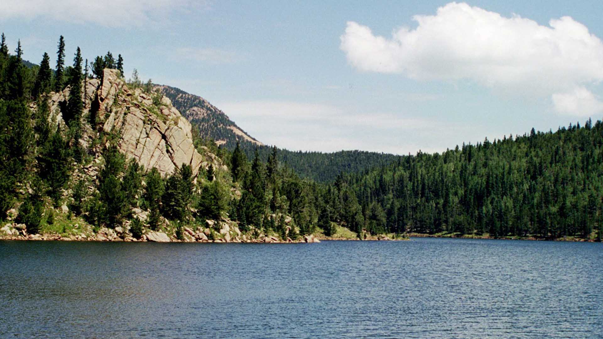 Rosemont Reservoir photographed under sunny skies. It is an alpine lake surrounded by rock cliffs and pine trees.