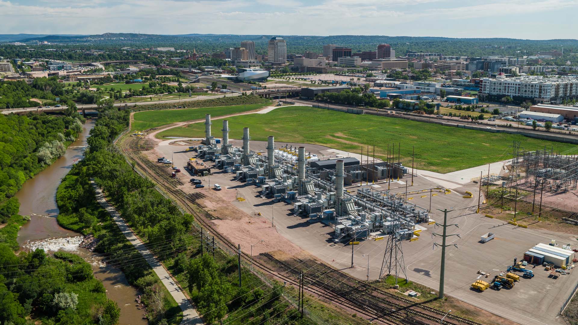 An aerial photo of the old site of the Martin Drake Power Plant with six new natural gas generating units in the foreground next to a green field. Downtown Colorado Springs is visible in the distance.