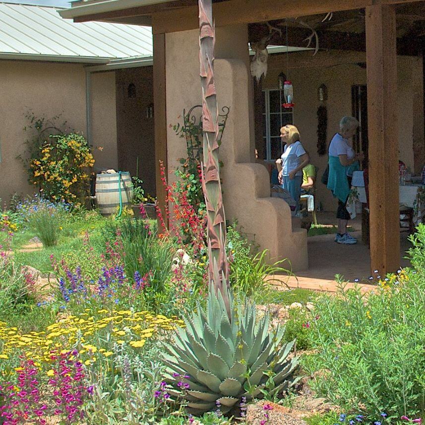 A vibrant garden with colorful flowers and a large agave plant, next to an adobe-style building with a wooden pergola.